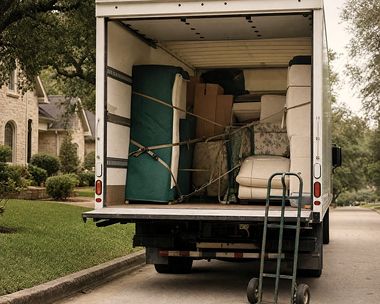 The open tailgate of a box truck loaded with moving-blanket-wrapped furniture, a dolly leaning against the bumper.